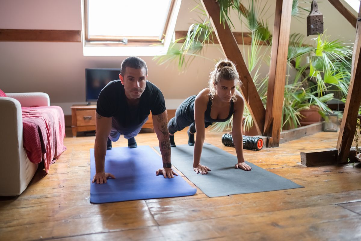 Casal em roupa esportiva fazendo flexões em colchonetes na sala de casa.